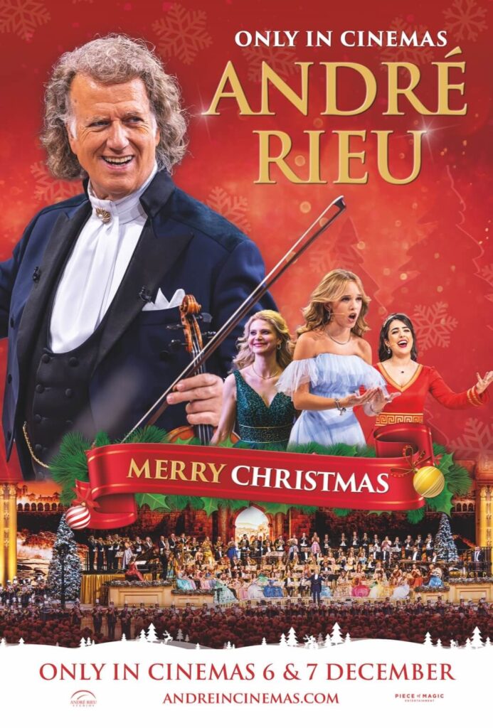 André Rieu holds a violin and stands in front of a red background as dancers do a waltz in his Christmas show
