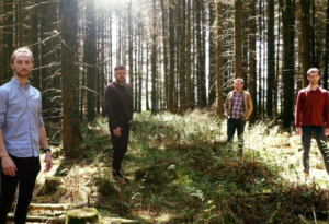 Four people stand in a forest with shafts of light coming through the trees