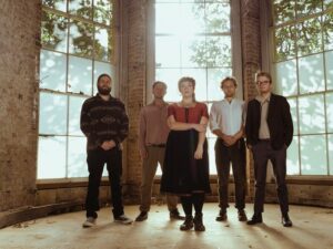 Five members of Around About Dusk stand by a large, sunlit window